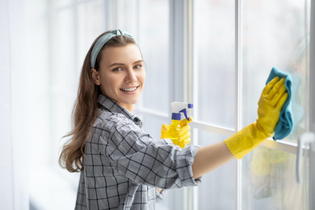 Smiling young woman with detergent and rag cleaning window glass. Professional sanitary service